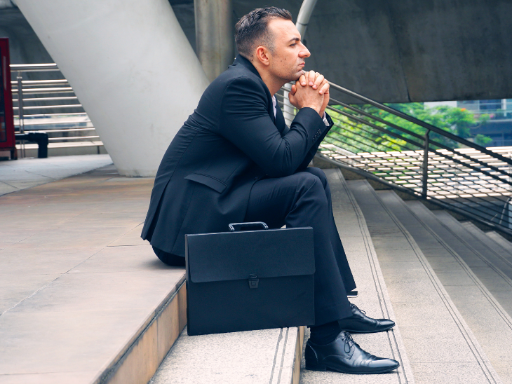 Man with concerned face, in suit, and with a briefcase sits on stairs.
